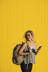 Lachende junge Frau mit Rucksack und Mobiltelefon vor gelbem Hintergrund - DAMF00144