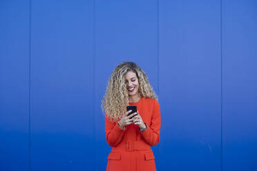 Porträt einer jungen Frau in einem roten Kleid, die ein Mobiltelefon vor einem blauen Hintergrund benutzt - DAMF00152