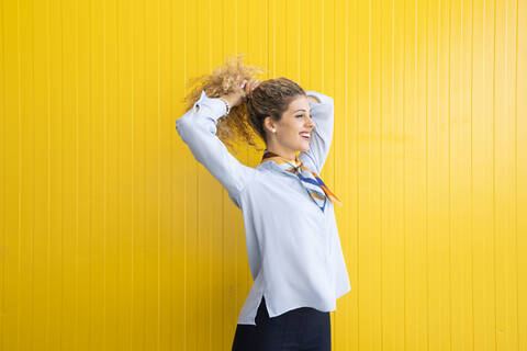 Lächelnde junge Frau, die ihre Haare vor einem gelben Hintergrund bindet, lizenzfreies Stockfoto