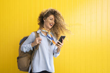 Lachende junge Frau mit Rucksack vor gelbem Hintergrund, die auf ihr Mobiltelefon schaut - DAMF00164