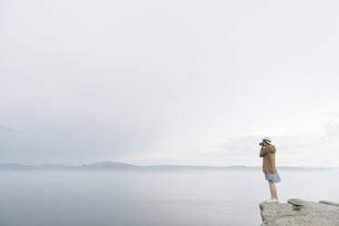 Frau mit Kamera, die auf einem Felsen steht und das Meer fotografiert, Rückansicht - AHSF00877