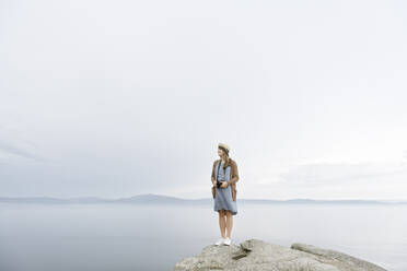 Woman with camera standing on rock, looking at the sea - AHSF00878