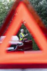 Nahaufnahme eines Warndreiecks vor dem kaputten Auto eines älteren Mannes auf einer Landstraße - AFVF04052