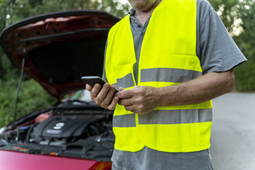 Close up of a senior man standing at his broken car wearing a safety vest and using his smartphone - AFVF04055