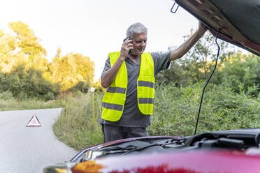 Ein älterer Mann steht an seinem kaputten Auto, trägt eine Sicherheitsweste und benutzt sein Smartphone - AFVF04056