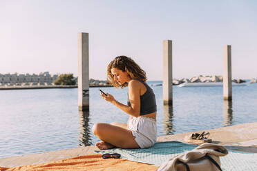 Young woman sitting on a pier at the sea using smartphone - MOSF00047