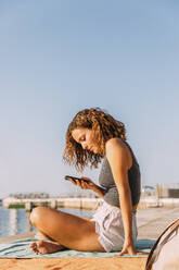 Junge Frau sitzt auf einem Pier am Meer und benutzt ihr Smartphone - MOSF00050