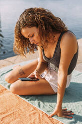 Junge Frau sitzt auf einem Pier am Meer und benutzt ihr Smartphone - MOSF00051
