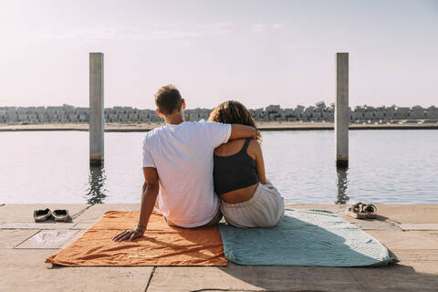 Rückansicht eines jungen Paares, das auf einem Pier am Meer sitzt, lizenzfreies Stockfoto