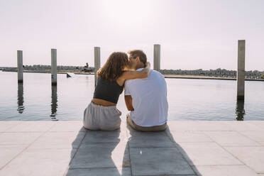 Rückansicht eines jungen Paares, das auf einem Pier am Meer sitzt und sich küsst - MOSF00062