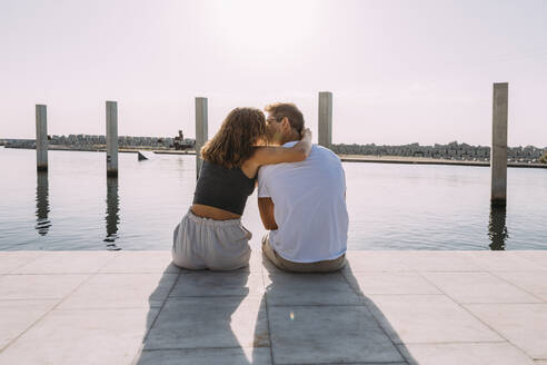 Rear view of young couple sitting on a pier at the sea kissing - MOSF00062