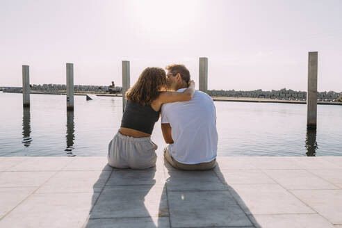 Rückansicht eines jungen Paares, das auf einem Pier am Meer sitzt und sich küsst - MOSF00062