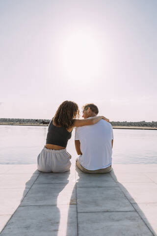 Rückansicht eines jungen Paares, das auf einem Pier am Meer sitzt, lizenzfreies Stockfoto