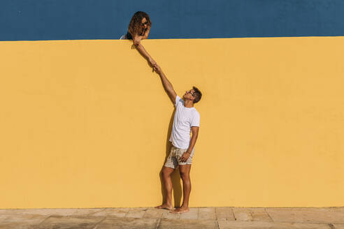 Young man and woman behind a yellow wall holding hands - MOSF00070