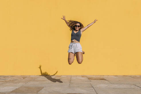 Happy young woman jumping in front of a yellow wall - MOSF00077