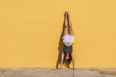 Young woman doing a handstand in front of a yellow wall - MOSF00078