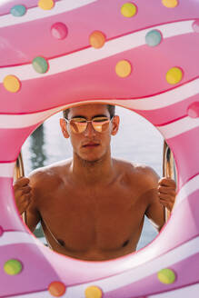 Portrait of young man behind inflatable float in donut shape - MOSF00094
