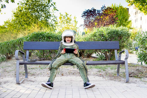 Junge in einem Raumanzug, der auf einer Bank sitzt und ein Tablet benutzt - CJMF00123