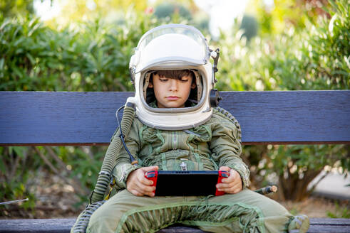 Boy wearing a space suit and sitting on a bench, playing alone with video games - CJMF00124