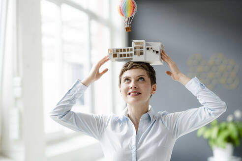 Smiling businesswoman carrying architectural model on her head with hot-air balloon floating above - KNSF06744