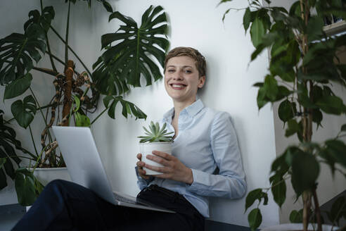 Porträt einer Geschäftsfrau mit Laptop auf dem Boden sitzend, umgeben von Pflanzen - KNSF06796