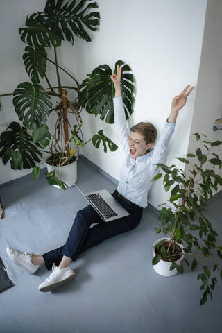 Jubelnde Geschäftsfrau mit Laptop auf dem Boden sitzend, umgeben von Pflanzen, lizenzfreies Stockfoto