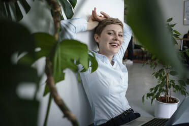 Portrait of a businesswoman with laptop sitting on the floor surrounded by plants - KNSF06798