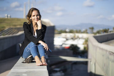 Portrait of confident businesswoman sitting on a wall with smartphone and earphones - GIOF07190