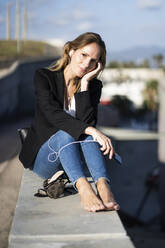 Portrait of confident businesswoman sitting on a wall with smartphone and earphones - GIOF07191