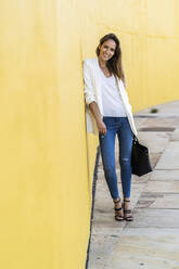 Porträt einer lächelnden Frau, die an einer gelben Wand steht und eine Handtasche hält - GIOF07210
