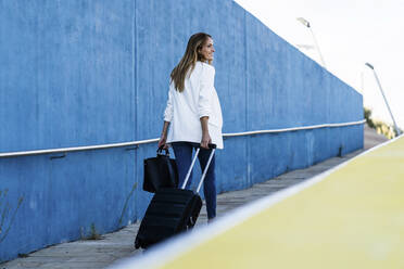 Businesswoman with baggage walking along blue wall - GIOF07214