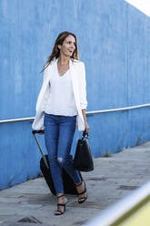 Glückliche Geschäftsfrau mit Gepäck geht an einer blauen Wand entlang - GIOF07216