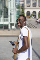 Smiling young man using smartphone and ear phones - CJMF00142