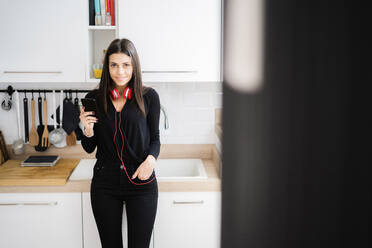 Portrait of smiling young woman with smartphone and headphones standing in her kitchen - GIOF07221