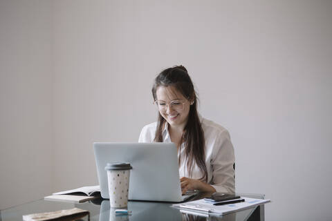 Lächelnde junge Frau arbeitet an einem Tisch im Büro und benutzt einen Laptop, lizenzfreies Stockfoto