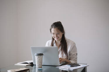Lächelnde junge Frau arbeitet an einem Tisch im Büro und benutzt einen Laptop - ALBF01180