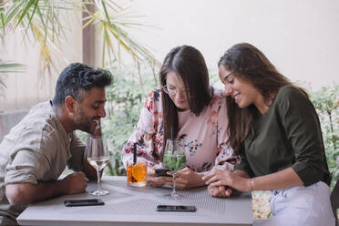 Lächelnde Freunde sitzen am Tisch mit Getränken und schauen auf ihr Smartphone - ALBF01200