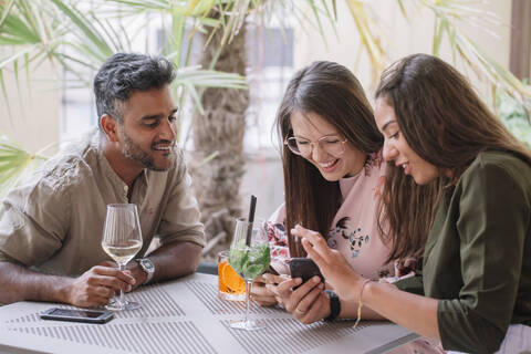 Glückliche Freunde sitzen am Tisch mit Getränken und schauen auf ihr Smartphone, lizenzfreies Stockfoto