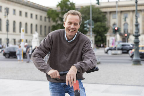 Porträt eines glücklichen älteren Mannes, der sich in der Stadt auf einen Elektroroller stützt, Berlin, Deutschland - WPEF02052