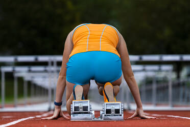 Germany, Baden Wurttemberg, Winterbach, Rear view of female hurdler kneeling at track starting block - STSF02297