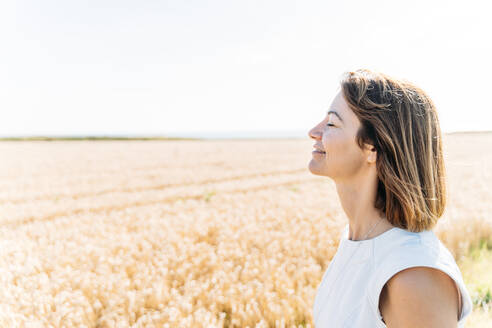 Frau mit geschlossenen Augen genießt den sonnigen Tag auf einem goldenen Feld. - CAVF65206