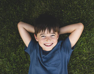 Overhead close up of happy boy laying on grass with arms behind head. - CAVF65240