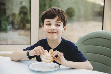 Porträt eines Jungen, der einen Sahne-Donut mit Sahne auf seiner Wange genießt - CAVF65301