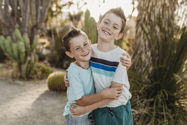 Cheerful brothers embracing and smiling in sunny cactus garden - CAVF65312