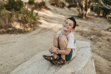 Porträt eines älteren Jungen, der auf einem Felsen sitzt und im Kaktusgarten lächelt - CAVF65321