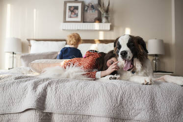 Kinder liegen mit einem großen Hund auf dem Bett, während das Mädchen versucht, die Zunge des Hundes zu berühren - CAVF65328