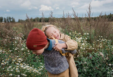 Bruder trägt seine Schwester lachend durch ein Wildblumenfeld - CAVF65339