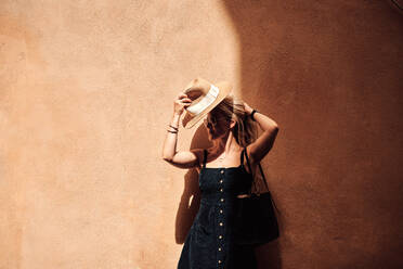 Stilvolle Frau, die einen Hut vor einer bemalten Wand aufsetzt - CAVF65351