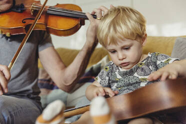 Porträt eines Kleinkindes, das die Ukulele testet, während sein Vater Geige spielt - MFF04892