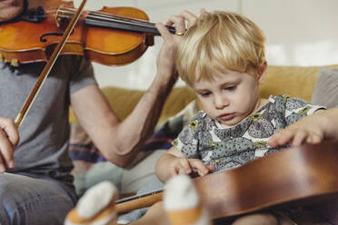 Portrait of toddler testing ukulele while his father playing violin - MFF04892